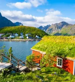 Rød norsk hytte med grønt græstag ved vandet, omgivet af bjerge og små huse, Lofoten, Reine, Norge. Reine 1-1. Togrejser Europa