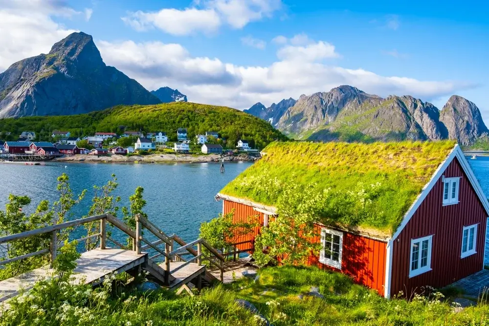 Rød norsk hytte med grønt græstag ved vandet, omgivet af bjerge og små huse, Lofoten, Reine, Norge. Reine 1-1. Togrejser Europa